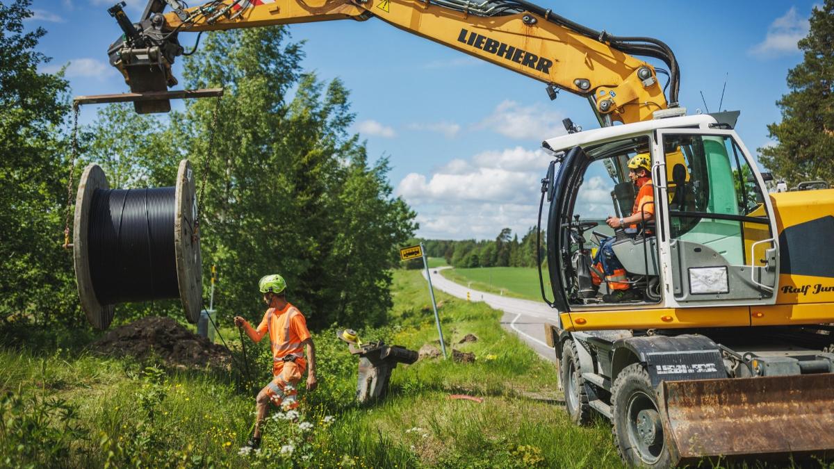 Kaivinkone nostaa valokuitukaapelirullaa kesäisessä maalaismaisemassa.