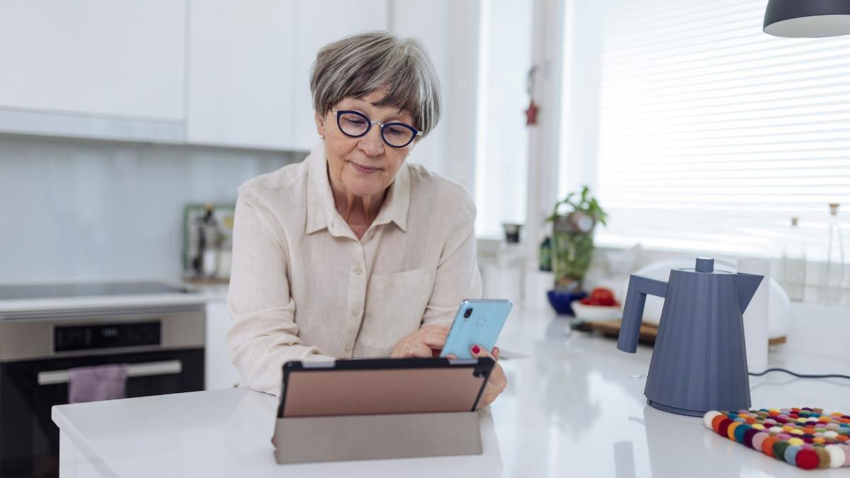 Ihminen istuu keittiön pöydän äärellä puhelin käsissään ja katsoo tablettia.