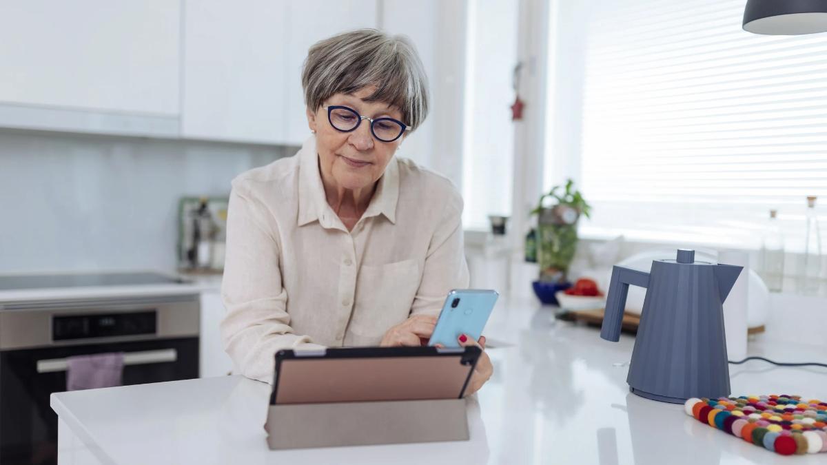 Seniori keittiön pöydän äärellä käyttää tablettia ja puhelinta.
