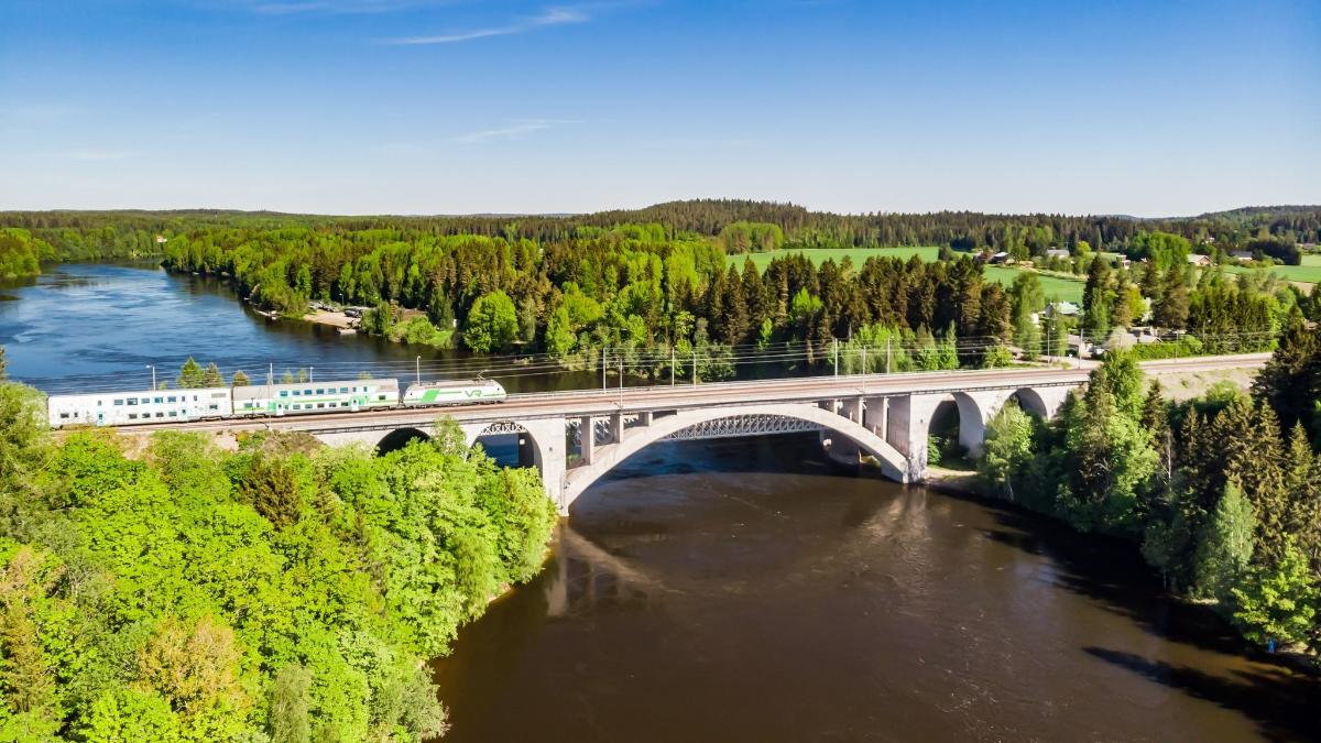 Kuva kesäisestä maisemasta Kymijoelta. Kuvassa rautatiesilta jota ylittää juna. Taka-alalla puita.