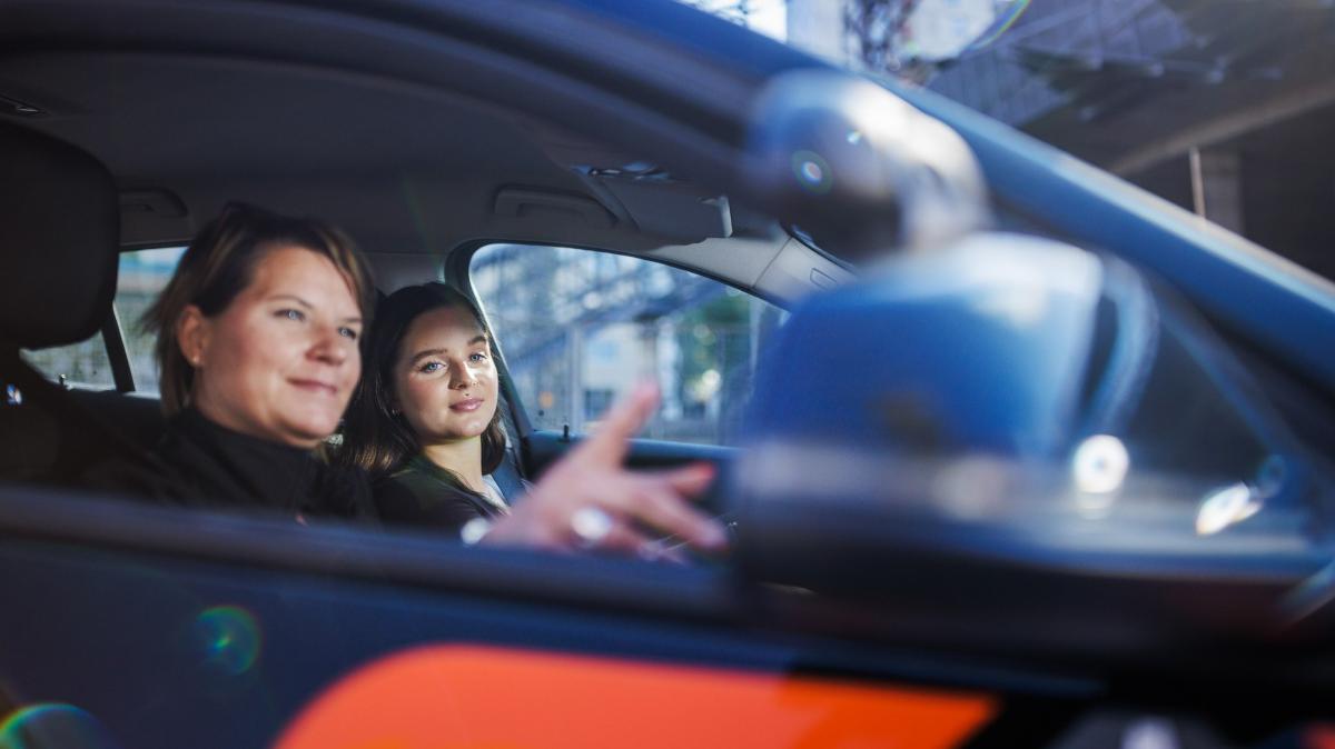 Nuori nainen ajo-opetuksessa autossa. Ajo-opettaja osoittaa mustan auton sivupeiliä.