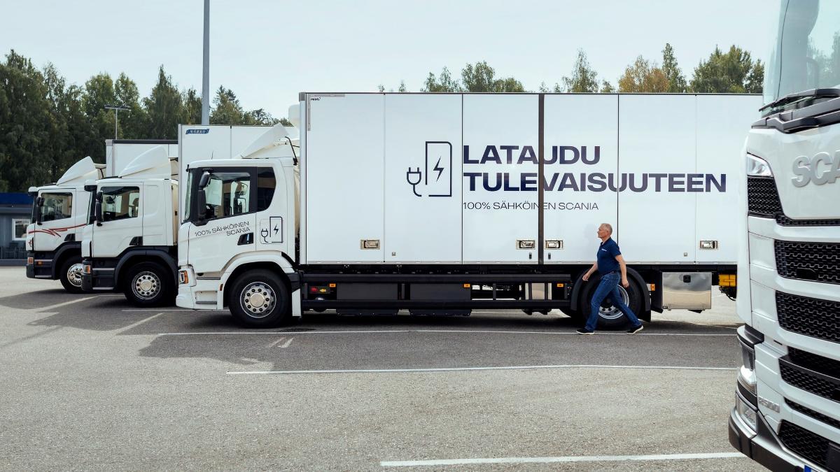 Electric lorries side by side, driver walking beside one of them. Text “Lataudu tulevaisuuteen” (Recharge for the future) written on the side wall of an electric lorry and a drawing of a power socket and battery beside it.