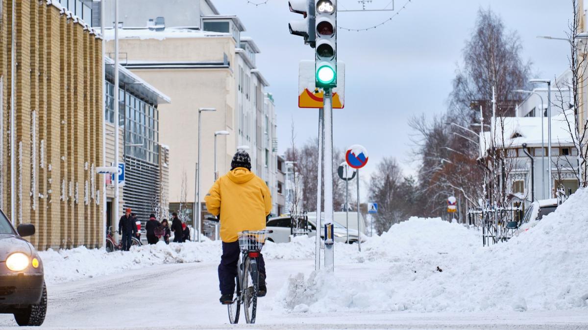 Keltatakkinen pyöräilijä ylittää katua lumisessa maisemassa