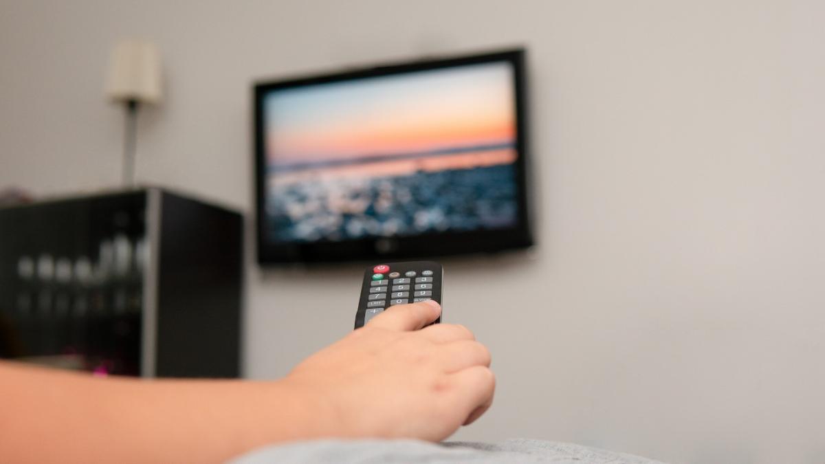 Kuvassa etualalla sohvan reunaan nojaava käsi, jossa on kaukosäädin. Taustalla näkyy seinässä oleva televisioruutu ja kirjahylly.