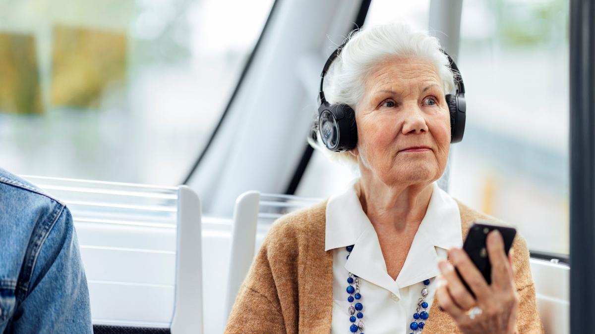 Iäkäs nainen istuu bussissa kuulokkeet päässä, puhelin kädessä.