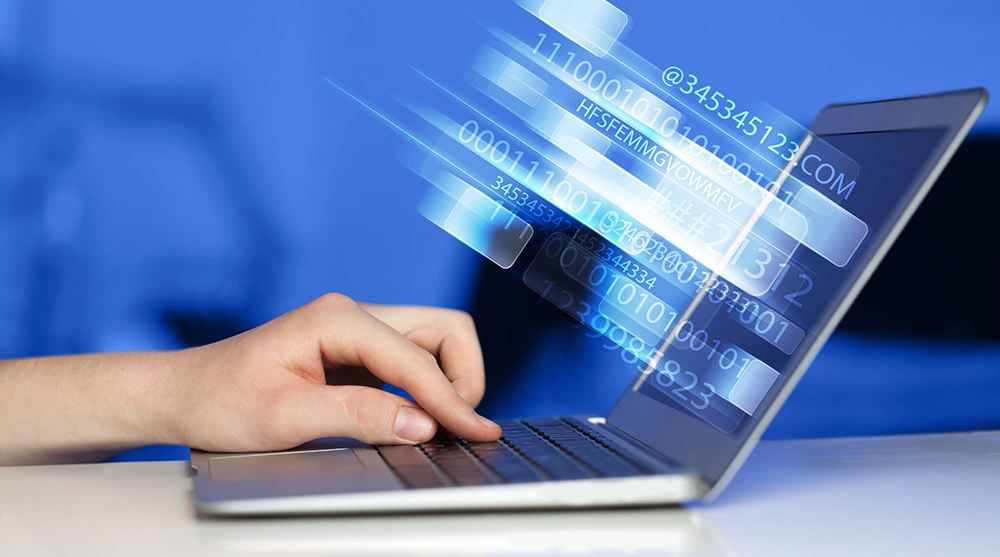 Kuvassa kädet näppäilevät kannettavaa tietokonetta ja merkkijonot symboloivat verkon välityksellä lähtevää tietoa